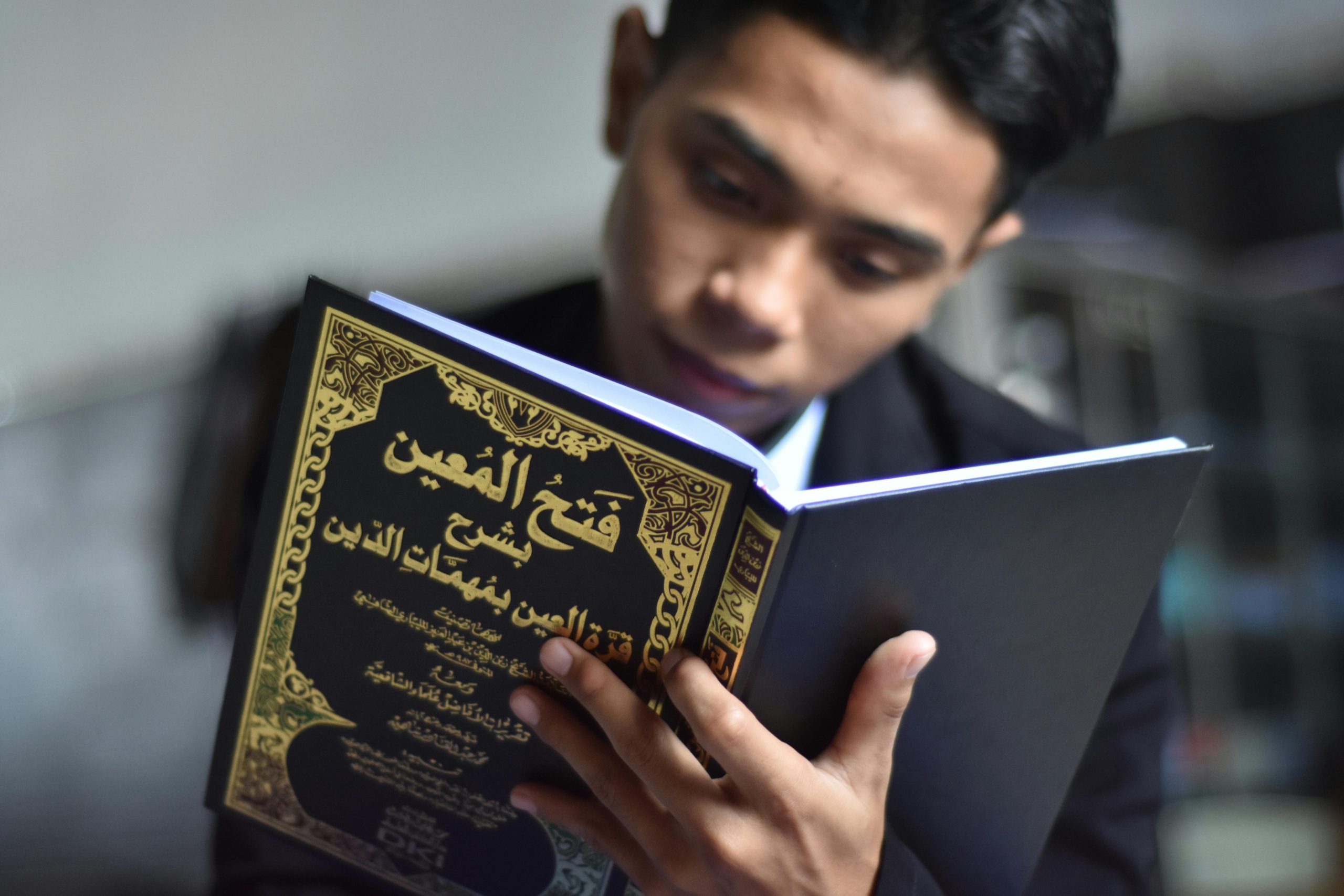 A young man intently reads an Islamic book indoors, showcasing cultural engagement.